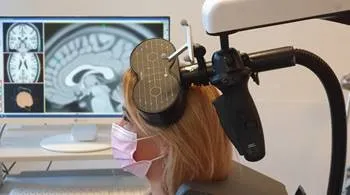 une personne est vue de profil, un appareil en forme de huit posée sur le front. En arrière plan, un écran avec une image d'IRM de cerveau
