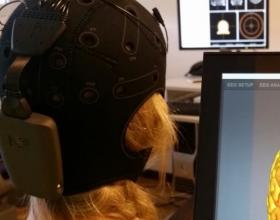 Une personne portant un bonnet en néoprène avec un petit boitier fixé à l'arrière, regardant un écran sur lequel un cerveau en 3D est affiché